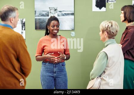Portrait of cheerful young Black female creator standing in front of art gallery exhibition visitors speaking about photos Stock Photo