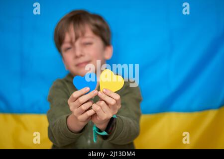 Boy hands holding yellow-blue paper hearts on Ukrainian flag background. Kids support Ukraine, stop war, stay with Ukraine hashtag Stock Photo