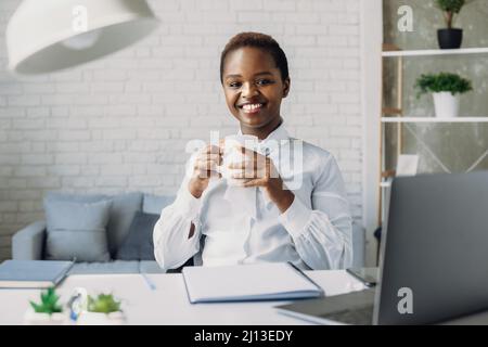 Indoor portrait of pleased african american woman looking at camera while posing in office during coffee break. Businesswoman portrait. Stylish Stock Photo