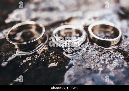 Three wedding rings of gold, platinum, silver and precious stones. Close-up portrait on the surface of a wet stone. Engagement ring. Stock Photo