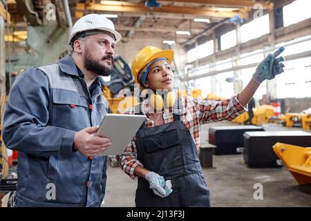 Young Hispanic female builder in workwear pointing at new equipment while discussing its characteristics with male colleague Stock Photo