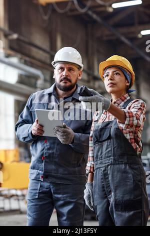 Engineers of large contemporary industrial plant looking at huge machine while female regulating its work and man using tablet Stock Photo