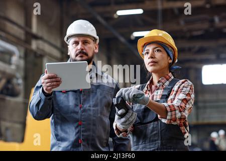 Two contemporary engineers with tablet and remote control looking at huge industrial machine while female regulating its work Stock Photo