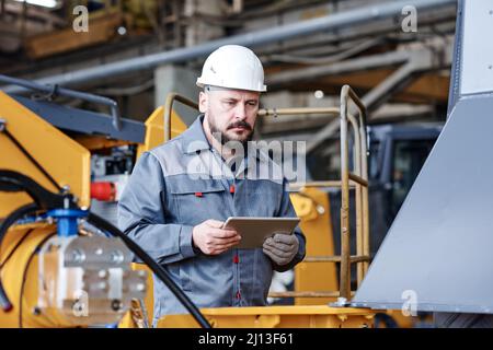 Mature serious engineer or quality control manager of industrial plant with tablet standing in front of huge industrial machine Stock Photo