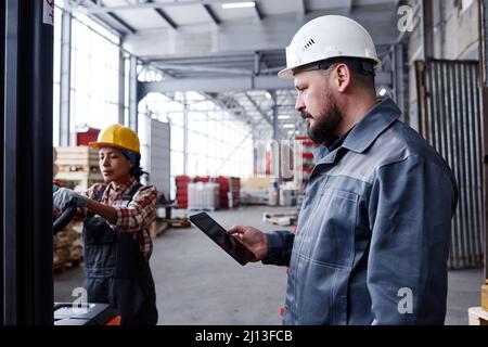 Bearded foreman or picker with tablet checking online orders in warehouse while young female worker pushing cart Stock Photo