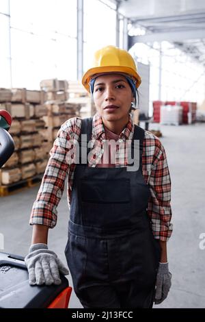 Young serious female engineer in workwear standing by cart with package in front of camera in distribution warehouse Stock Photo