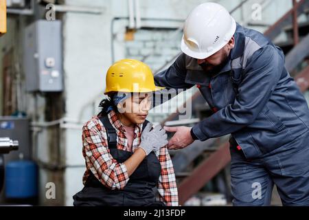 Foreman in hardhat and workwear standing by female engineer with contusion touching her shoulder against machine Stock Photo