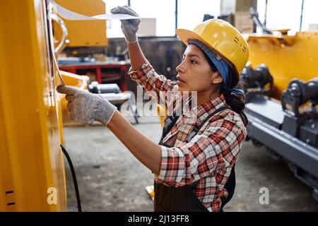 Young Hispanic woman in coveralls and safety helmet looking at working plan on paper hanging on side of industrial machine Stock Photo