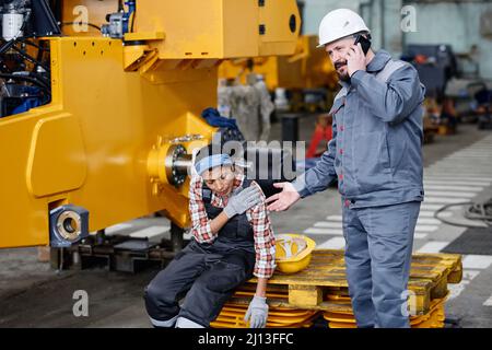 Mature worker of warehouse in workwear calling ambulance for his female colleague with injury or contusion in her shoulder Stock Photo