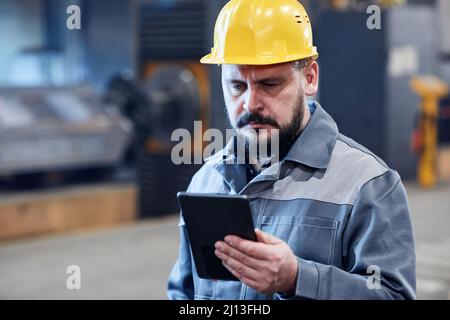 Mature bearded male worker of industrial plant in boilersuit and hardhat looking through data on screen of digital tablet Stock Photo