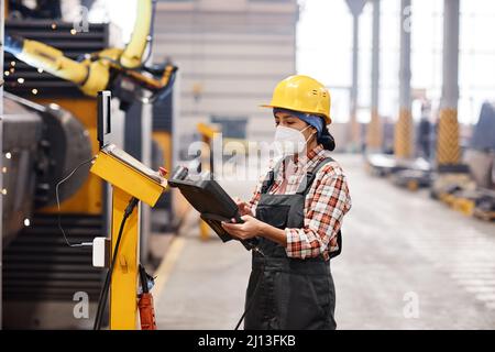 Young female technician in protective workwear using remote switch to control industrial machine while standing in large workshop Stock Photo