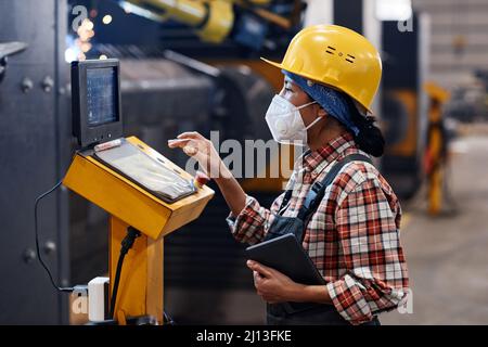 Young woman in protective helmet, respirator and coveralls pointing at document while looking at data on screen during work Stock Photo