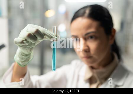 Gloved hand of young female scientist in lab coat holding flask with blue liquid while carrying out scientific experiment in laboratory