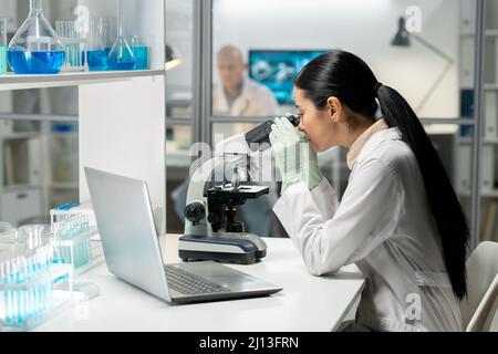 Side view of young female chemical researcher working with microscope while studying new substances by workplace in laboratory Stock Photo