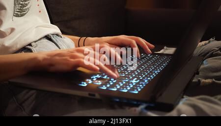 Close up woman hands working typing on keyboard laptop computer at dark room sitting on couch. Freelancer using laptop working from home brightening k Stock Photo