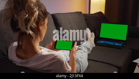 Over the shoulder view woman lying on a sofa using phone and laptop with Green Mock-up Screen Chroma key watching content. Horizontal phone Green Scre Stock Photo