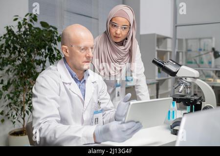 Two young contemporary virologists discussing online data in laboratory whilemature male scientist pointing at tablet screen Stock Photo