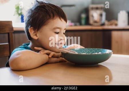 Caucasian boy is bored at the table with his head on hands, does not want to eat oatmeal porridge. Happy face. Stock Photo
