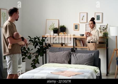 Young woman with cup of coffee standing by doubel bed in front of her husband holding sleeping baby son on hands Stock Photo