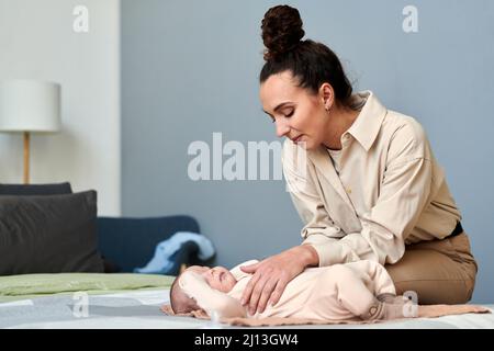 Young contemporary female in smart casualwear bending over her baby son lying on double bed while going to get him dressed Stock Photo