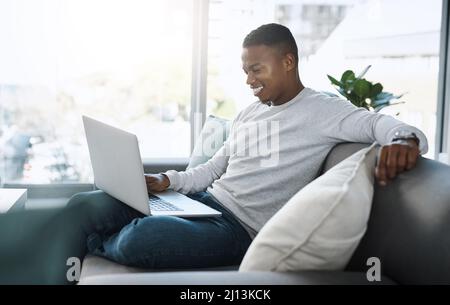 Where do I sign up for this content. Shot of a handsome young man using his laptop while sitting on a sofa at home. Stock Photo