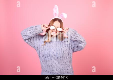 Beautiful girl in a blue sweater on a pink background. Woman with bunny ears, posing for easter, covering her eyes with easter eggs Stock Photo