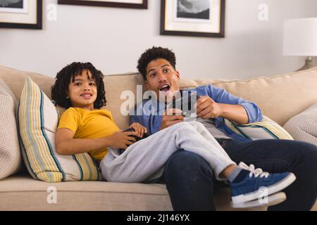 Cheerful hispanic father and son playing video games while sitting on sofa in living room Stock Photo