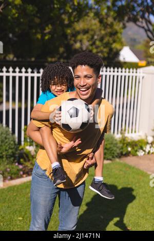 Portrait of happy hispanic father giving piggyback ride to son holding soccer ball at backyard Stock Photo
