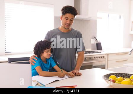 Hispanic man standing by son doing homework at dining table in kitchen Stock Photo