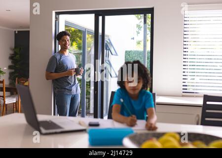 Smiling hispanic man standing on doorway looking at son doing homework in kitchen Stock Photo