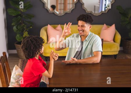 Smiling hispanic father teaching sign language to mute son while sitting at dining table Stock Photo