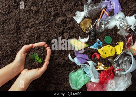 Top view of the hands that hold young sprout against the background of plastic debris on the ground, plastic problem concept Stock Photo