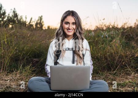 Young caucasian woman with a backpack on, using a laptop for teleworking in the middle of the field. Digital nomad. Stock Photo