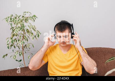 A young man in a yellow T-shirt talks on headphones, works from home and communicates with a client on the Internet. Room with houseplants. Concept of online education and homework, freelance. Stock Photo