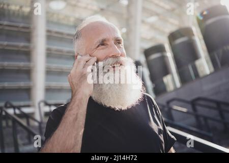 Photo of thoughtful handsome man pensioner dressed black clothes talking modern device outdoors city street Stock Photo