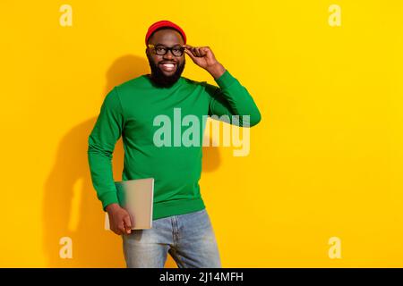 Portrait of attractive successful cheerful guy holding laptop touching specs isolated over bright yellow color background Stock Photo