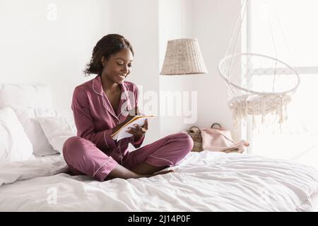 Black Female Taking Notes Writing In Notebook Sitting In Bedroom Stock Photo