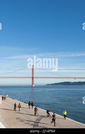 Joggers and walkers strolling along the Rio Tejo with the iconic suspended bridge (Ponte 25 de Abril), in the background - Lisbon, Portugal Stock Photo