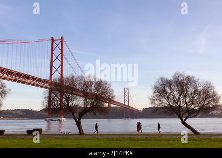 Joggers and walkers strolling along the Rio Tejo with the iconic suspended bridge (Ponte 25 de Abril), in the background - Lisbon, Portugal Stock Photo