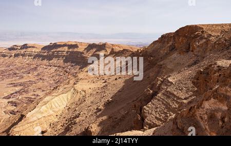 section of the south rim of the Makhtesh Katan Small Crater showing cliffs and colorful sandstone with the Jordan Rift Valley in the background Stock Photo