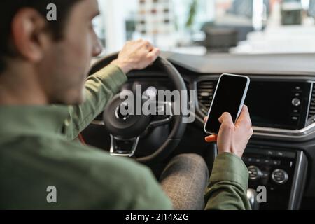Unrecognizable young man using smartphone, sitting in drivers seat of new car, buying vehicle at auto dealership, mockup Stock Photo
