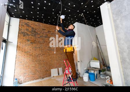 Full Length Portrait Of Electrician On Stepladder Installs Lighting To The Ceiling In Office Stock Photo