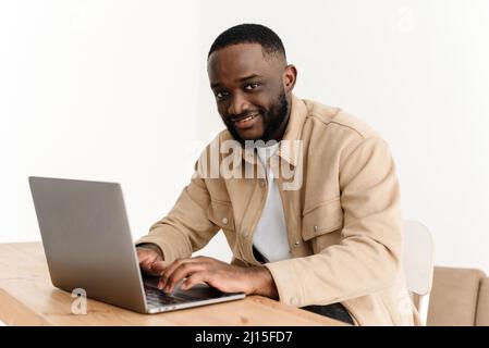 Portrait of smiling african american man looking at camera while sitting at table in home office and working on laptop. Happy black freelancer posing Stock Photo