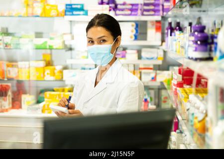 Portrait of female pharmacist in medical mask working at the cash register in pharmacy Stock Photo