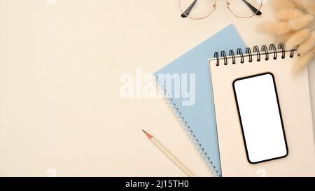 Overhead shot, Modern workspace with smartphone blank empty screen mockup on spiral notepad, eyeglasses, pencil and copy space on white background. Stock Photo