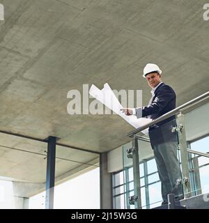 Going through the plans like the boss he is. Portrait of a cheerful professional male architect looking at the camera while holding blueprints inside Stock Photo