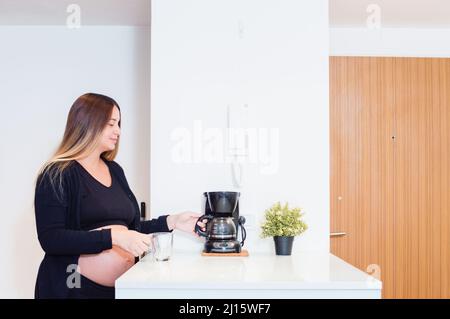 pregnant young caucasian woman in the kitchen of her apartment preparing breakfast and coffee for her husband, taking the pot of coffee from the coffe Stock Photo