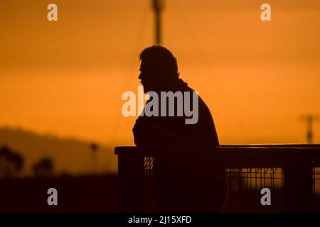 Silhouette of man with beard at sunset leaning on fence looking out at view Stock Photo