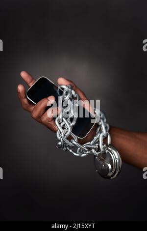 Tied to technology 247. Cropped shot of a mans phone tied to his hand with chains against a black background. Stock Photo