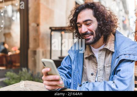 Positive bearded Hispanic man in casual clothes with curly hair smiling and watching video on smartphone while sitting at table in street restaurant Stock Photo